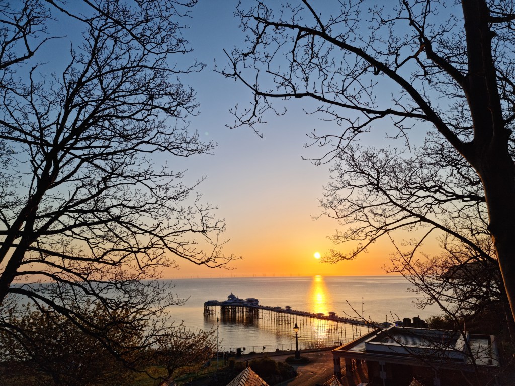 Sunrise view of Llandudno Pier from a path in Happy Valley Botanical Gardens, with warm morning light and coastal scenery