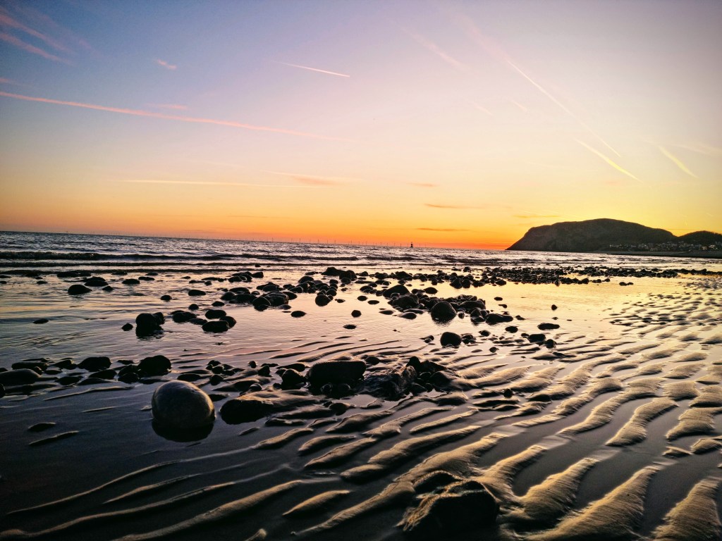 Sunrise over Little Orme with pink and orange hues reflected on wet sand at low tide, viewed from North Shore beach in Llandudno