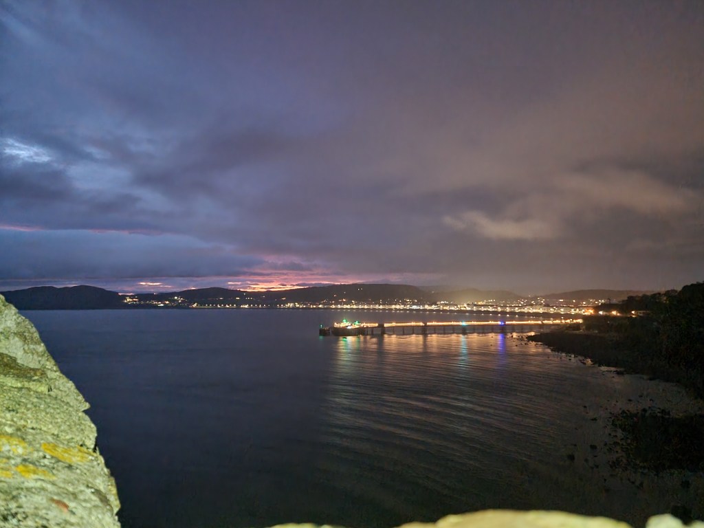 View of Llandudno Pier and town from the Great Orme road at dawn, with glowing lights and a dim, early sky.