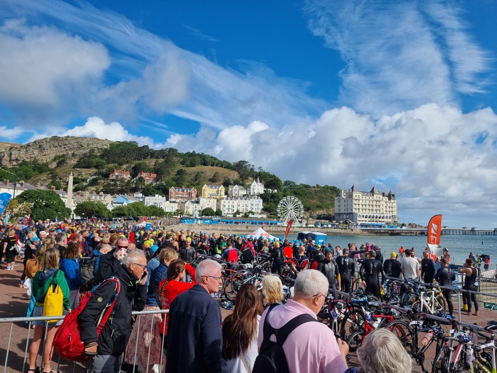 Participants and spectators gathered before the Llandudno Triathlon start with Min y Don, Llandudno Pier, and the seafront in the background.